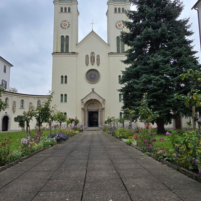 Eine weiße Kirche mit zwei Türmen, jeder mit einer Uhr, steht vor einem Garten mit Blumen. Die Kirche hat ein Kreuz auf der Spitze und ein Fenster in der Mitte.