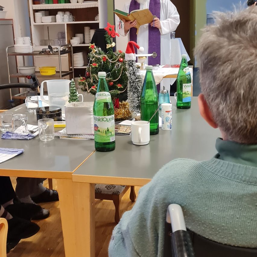 Ein Priester liest ein Buch an einem mit einem Weihnachtsbaum, grünen Flaschen und verschiedenen Gegenständen geschmückten Tisch. Gegenüber sitzt eine Person, und ein Regal mit Tassen und Schüsseln ist im Hintergrund.