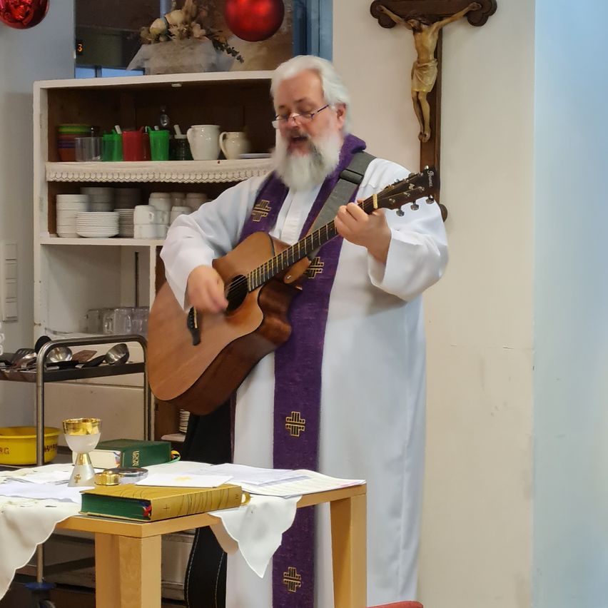 Ein Priester spielt Gitarre in einem Raum mit Tisch, Kelch und Kreuz an der Wand.