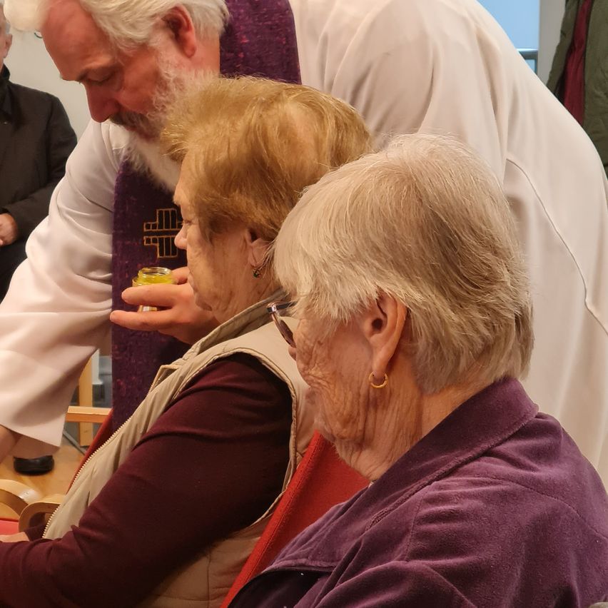 Ein Priester legt seine Hand auf den Kopf einer älteren Frau, die aus einem Becher trinkt. Eine weitere ältere Frau mit Brille sitzt in der Nähe.