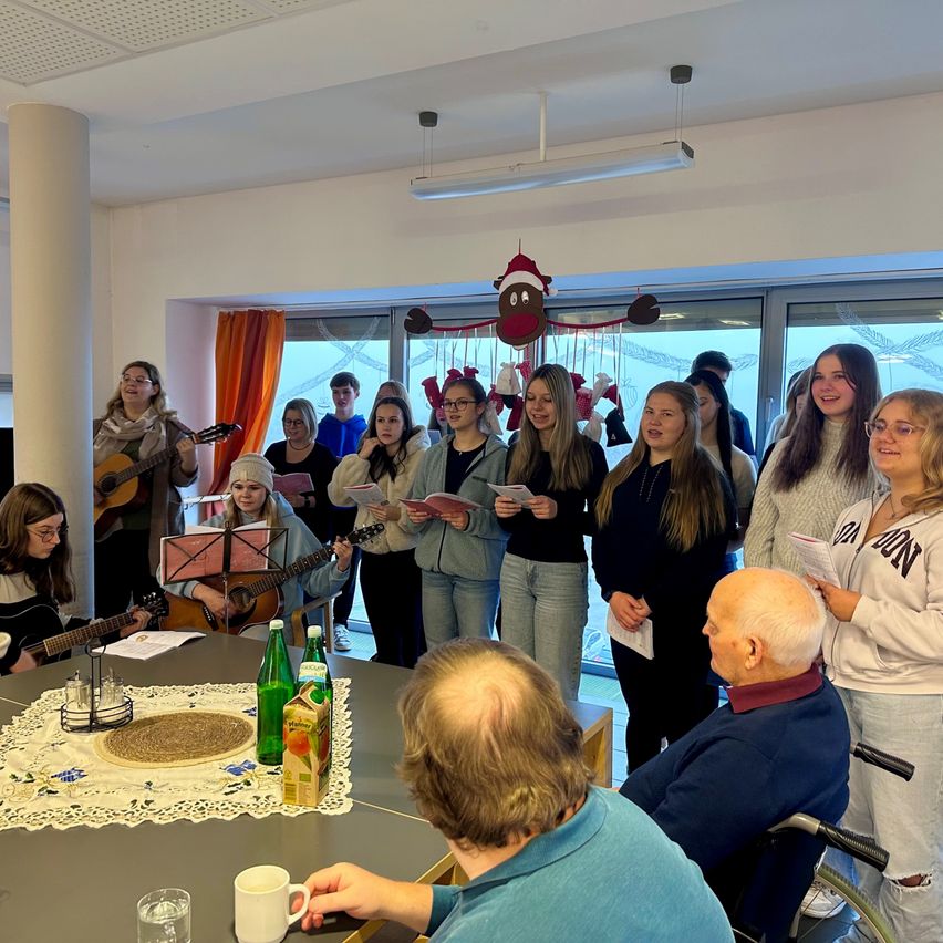 Eine Gruppe von Frauen und einige Männer versammeln sich um einen Tisch und singen gemeinsam mit Gitarren, während ein Mann im Rollstuhl aufmerksam zuhört. Der Raum hat eine festliche Atmosphäre mit Dekorationen.