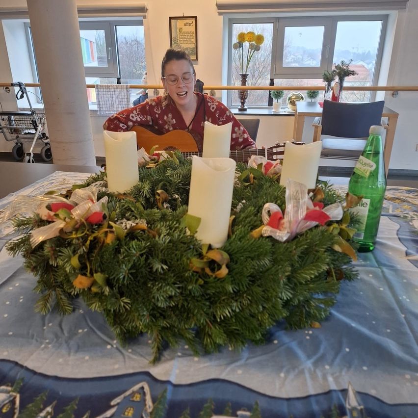 Eine Frau spielt Gitarre in einem Raum mit einem Kranz aus grünen Blättern und vier weißen Kerzen. Der Raum hat Fenster mit Blick nach draußen und einen dekorierten Tisch.