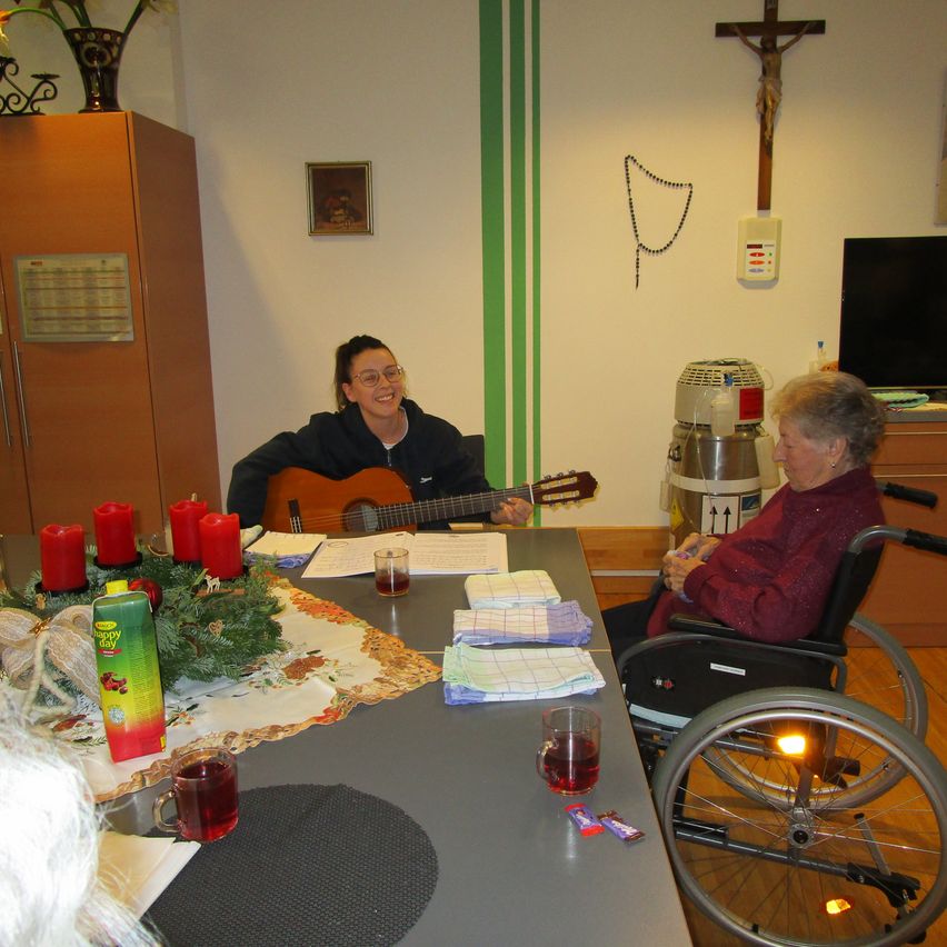 Eine Frau im Rollstuhl sitzt an einem Tisch mit Kerzen, Blumen und Servietten. Eine andere Frau spielt neben ihr Gitarre. Ein Kruzifix und eine Topfpflanze hängen an der Wand.