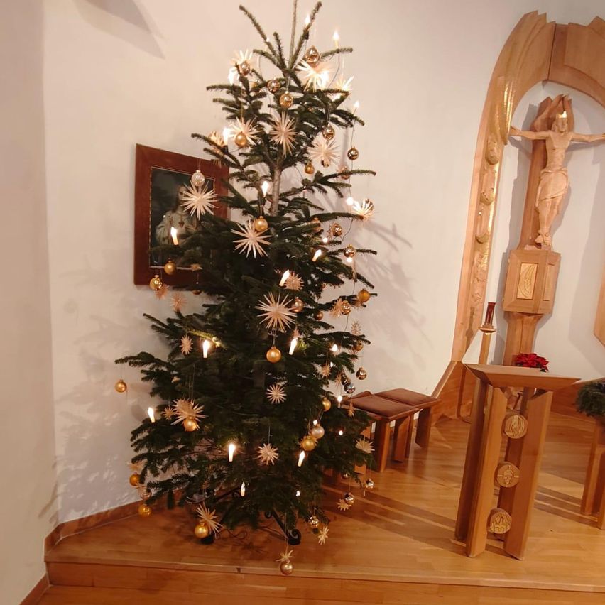 Ein Weihnachtsbaum ist mit Ornamenten und Kerzen geschmückt. Er steht in einem Raum mit einem Holzkreuz und einer Bank. Der Raum hat Holzböden und weiße Wände.