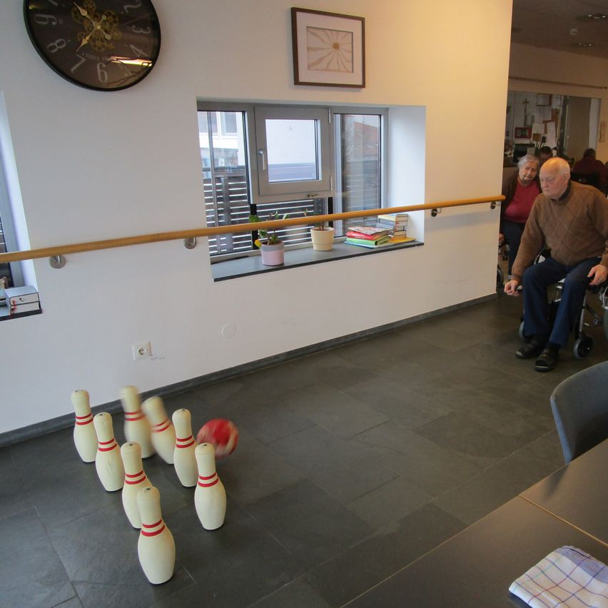 Ein Mann im Rollstuhl spielt drinnen Bowling, während andere zusehen. Eine Uhr und ein Bilderrahmen sind an der Wand darüber.