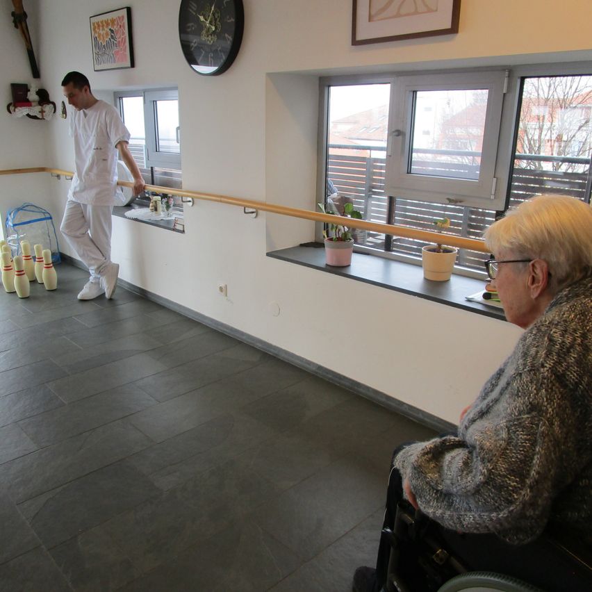 Ein Mann in Weiß geht an einem Handlauf in einem Raum mit einem Rollstuhl vorbei. Eine Frau sitzt im Rollstuhl und schaut zum Fenster.