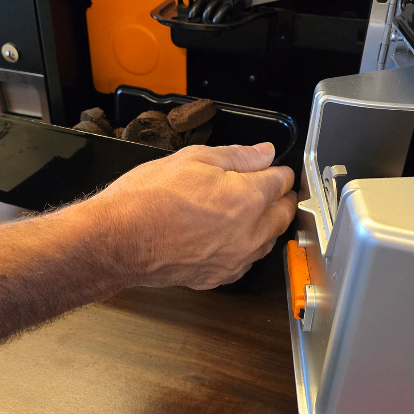 Eine Hand steckt eine orangefarbene Lasche in eine Metallbox, die mit Holzkohle gefüllt ist. Dahinter steht ein schwarzer Behälter mit mehr Holzkohle, und links steht ein orangefarbener Behälter.