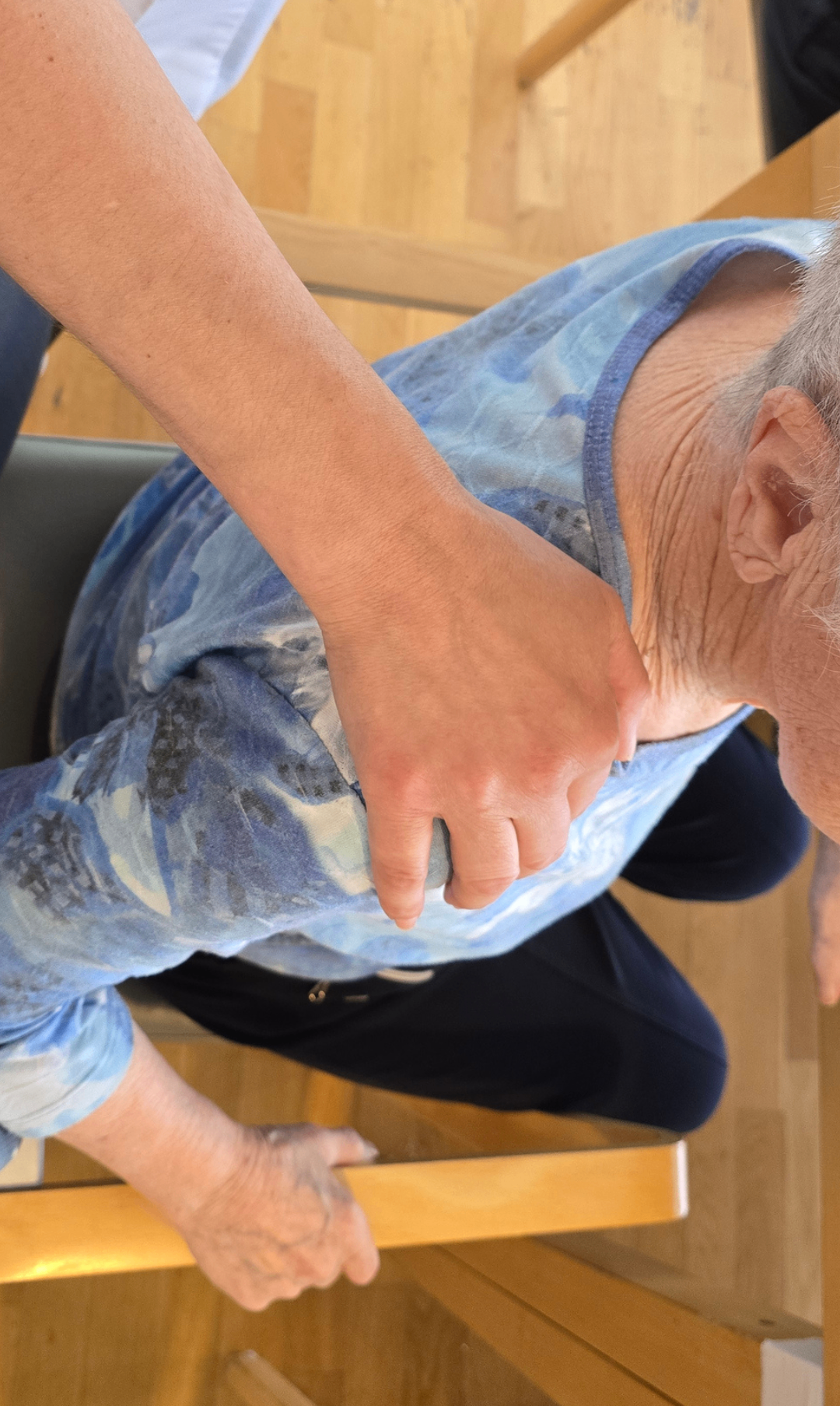 Eine ältere Person sitzt auf einem Stuhl mit der Hand einer anderen Person auf der Schulter. Die Hand ist positioniert, um Unterstützung oder Trost zu geben.