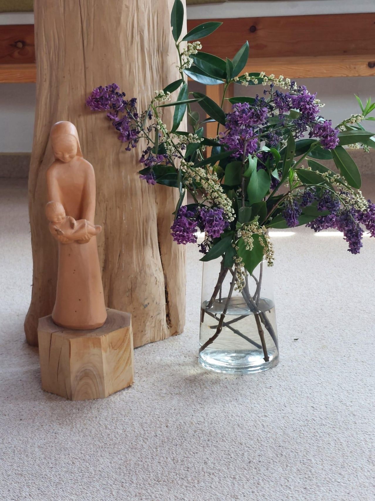 A wooden statue of a woman holding a baby stands on a wooden block next to a vase of purple flowers.