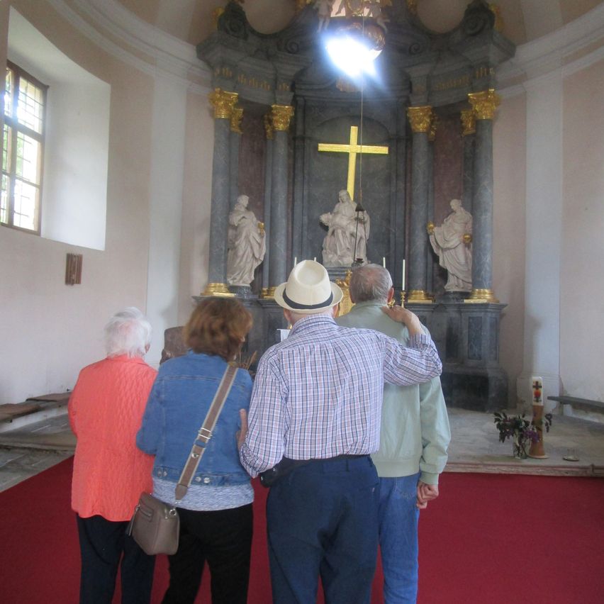 Inside a church, several people stand near an altar with a large cross and statues, on a red carpet.