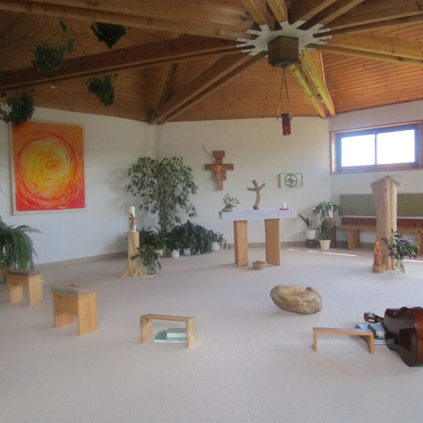 A serene chapel with wooden altar and benches. Potted plants and a large painting adorn the walls. A wooden ceiling beams and a window allow natural light.