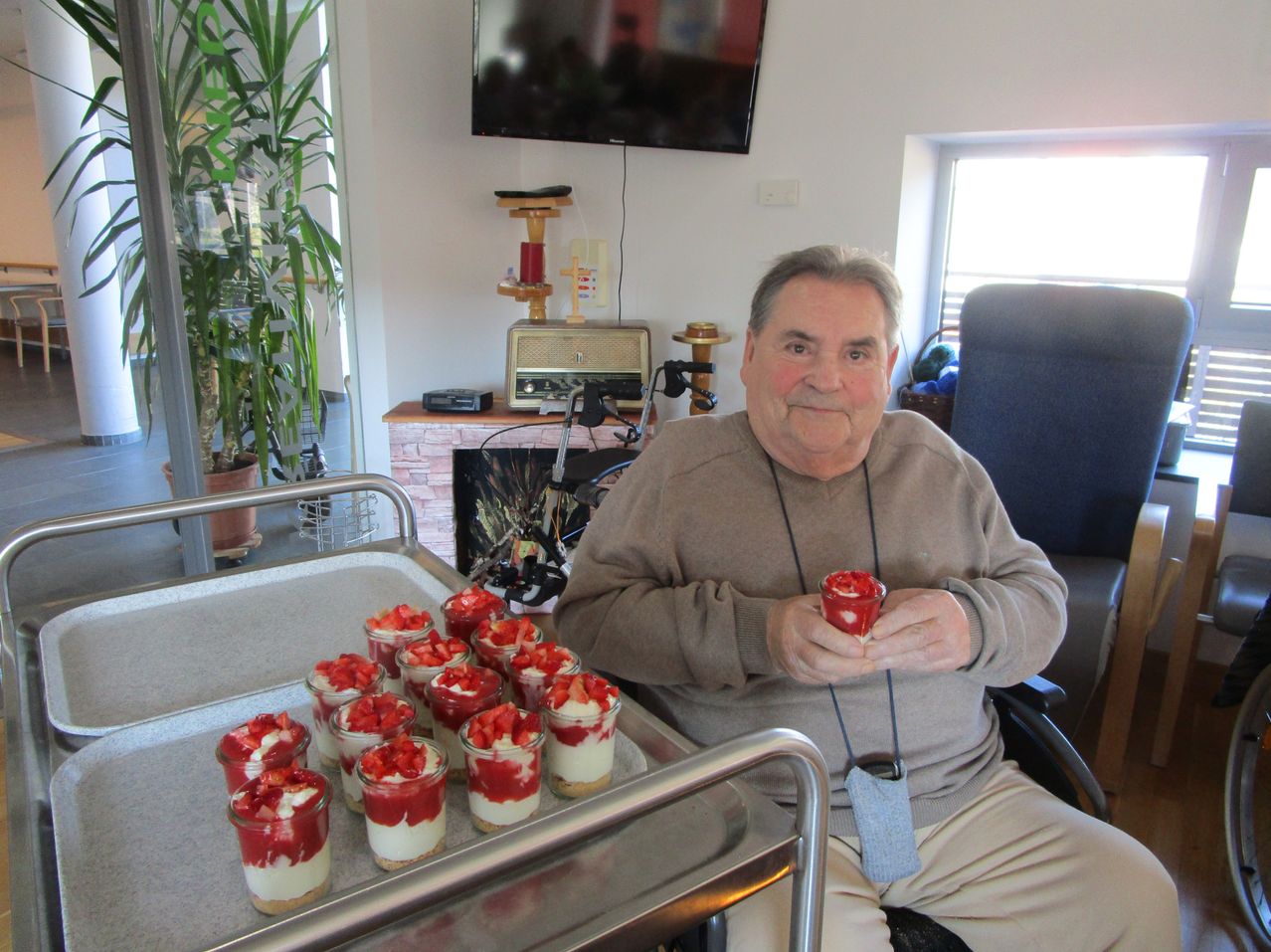 Ein älterer Mann im Rollstuhl sitzt in einem Raum mit einem Tablett mit Erdbeerdesserts vor ihm.