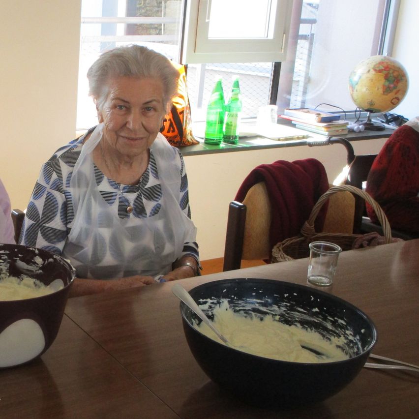 Eine ältere Frau sitzt an einem Tisch mit einer Schüssel Essen. Sie trägt eine Halskette und eine Schürze. Dahinter steht ein Regal mit Büchern und einer Globus.