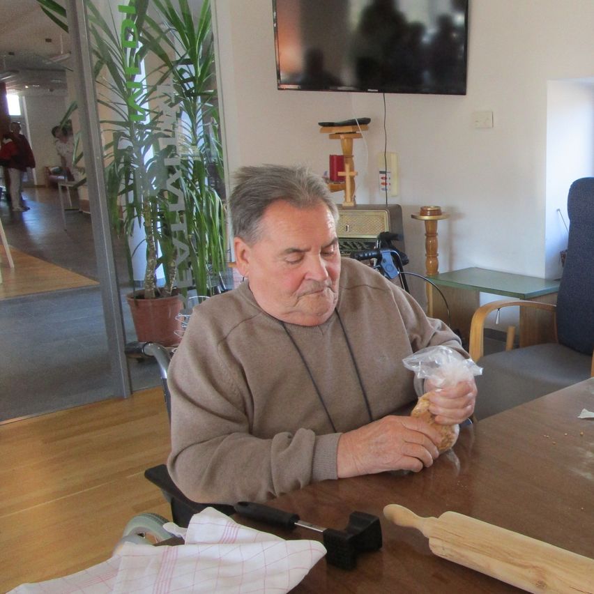 Ein älterer Mann sitzt an einem Tisch und hält eine Plastiktüte. Ihm zur Seite stehen Küchenutensilien, darunter ein Messer und ein Nudelholz. Dahinter steht eine Topfpflanze, ein montierter Fernseher und ein Stuhl. Im Hintergrund sind Personen zu sehen, eine von ihnen trägt einen Rucksack.