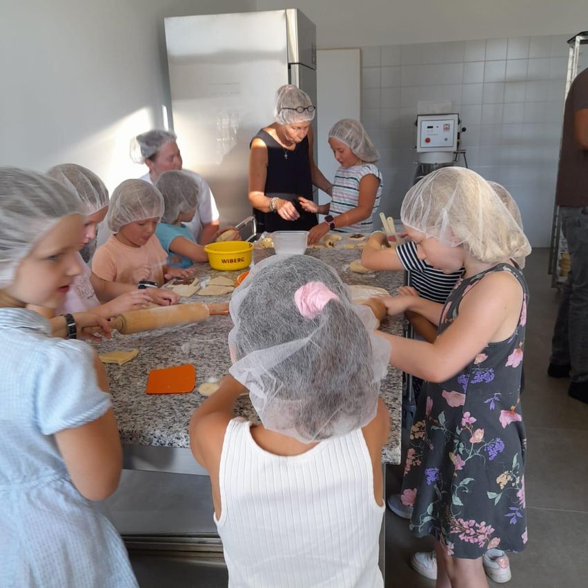 Eine Gruppe von Kindern mit Haarnetzen bereitet in einer Küche Teig zu, während eine erwachsene Frau beaufsichtigt.