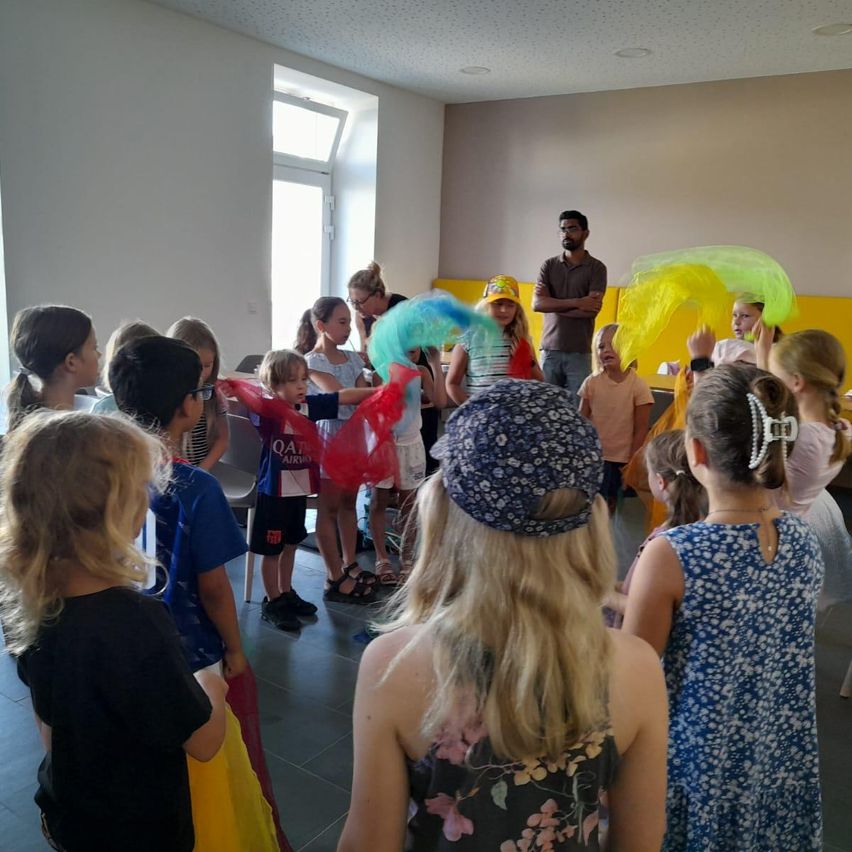 Eine Gruppe von Kindern und Erwachsenen versammelt sich in einem Raum, einige halten bunte Requisiten. Ein Mann steht im Hintergrund, und ein Fenster ist an der Wand zu sehen.