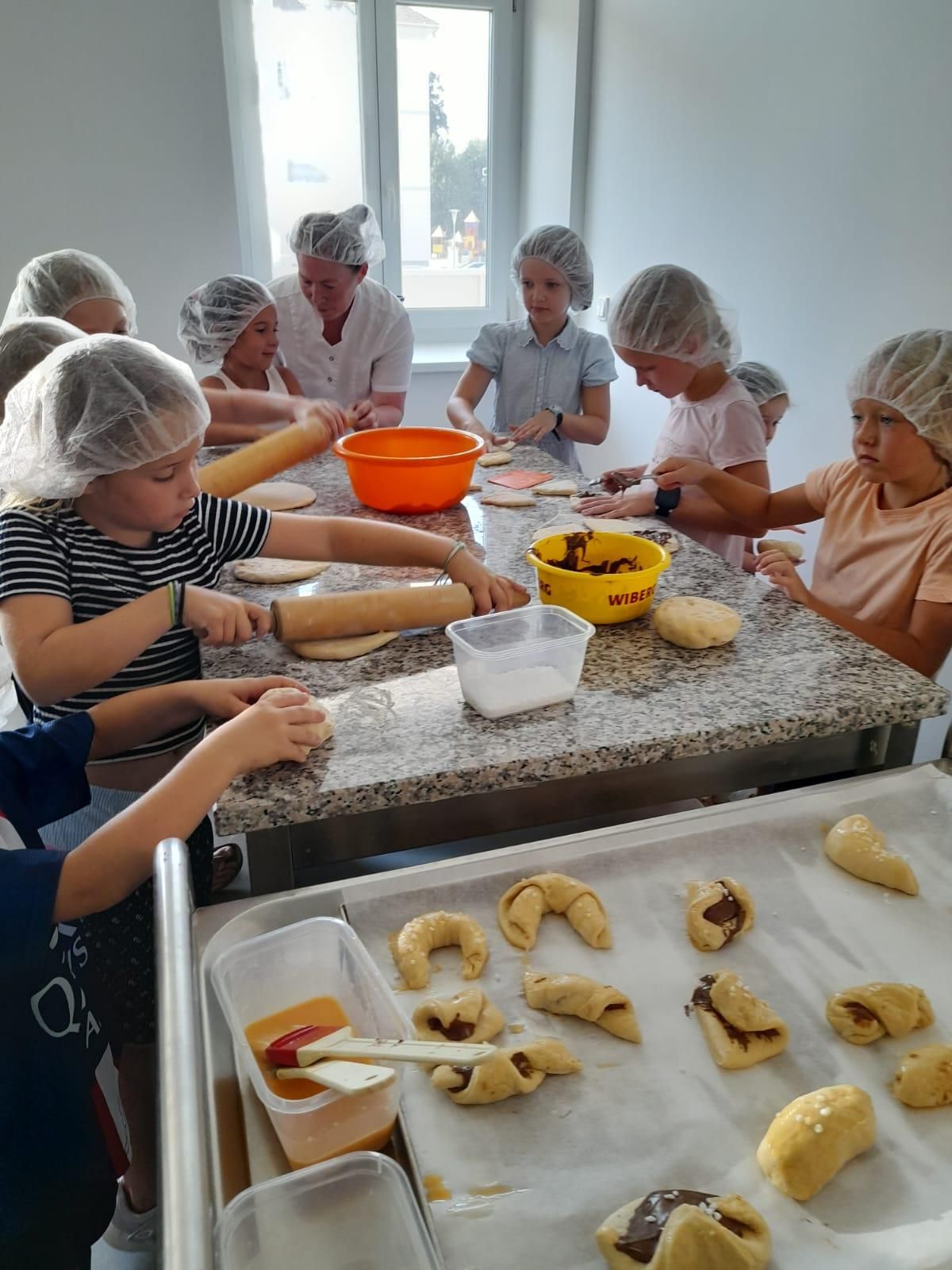 Eine Gruppe von Kindern mit Haarnetzen rollt Teig auf einem Tisch aus, wobei ein Kind einen Nudelholz verwendet. Schüsseln und Tabletts mit Teig stehen auf dem Tisch, und eine Frau steht hinter ihnen.