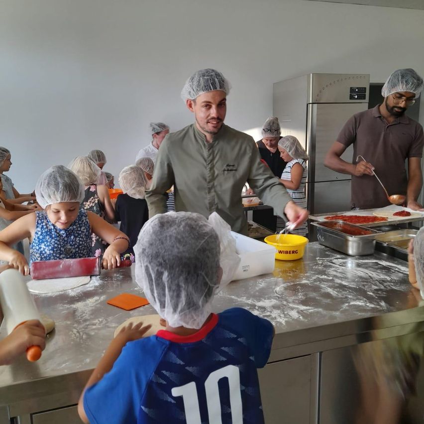 Eine Gruppe von Kindern und Erwachsenen befindet sich in einer Küche, trägt Haarnetze und Schürzen. Sie bereiten Essen zu, ein Mann in einem grünen Hemd steht an einem Tisch mit einer Schüssel und einem Löffel.