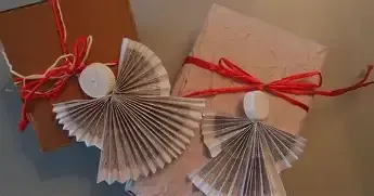 Zwei handgefertigte Engel-Dekorationen aus gefalteter Papier mit weißen Kerzen darauf, in braune Papier mit roten Bändern verpackt. Sie sind auf einer weißen Oberfläche platziert.