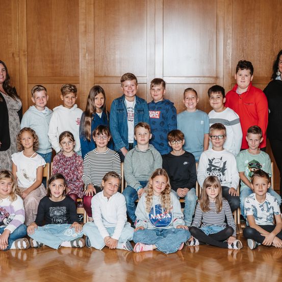 Eine Gruppe von Kindern und Erwachsenen steht und sitzt zusammen in einem Raum und lächelt für ein Gruppenfoto. Die Kinder tragen Freizeitkleidung, und die Erwachsenen sind im Hintergrund.