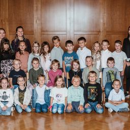 Eine Gruppenaufnahme von Kindern und zwei Erwachsenen, die vor einer Holzwand stehen, in zwei Reihen angeordnet, wobei die Erwachsenen hinten stehen.