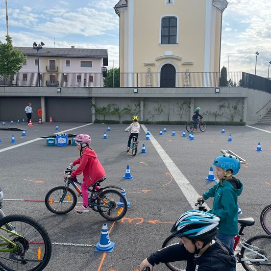 Kinder üben das Radfahren auf einem Parkplatz mit Hütchen. Eine Person steht bei einem Gebäude mit Statuen. Bäume und Straßenlaternen sind in der Nähe.