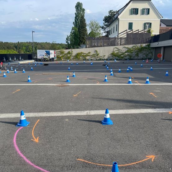 Ein Parkplatz mit zahlreichen blauen und weißen Kegel, die in einem kreisförmigen Muster angeordnet sind, einem rosa Kreis und Pfeilen auf der Straße. Ein Lieferwagen ist in der Nähe eines Zauns mit Bäumen im Hintergrund geparkt.