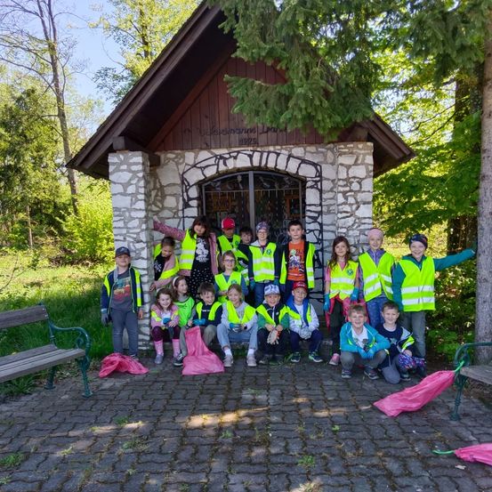 Eine Gruppe von Kindern und Erwachsenen in Warnwesten posiert für ein Foto in einer kleinen Kapelle mit Bänken und rosa Regenschirmen.