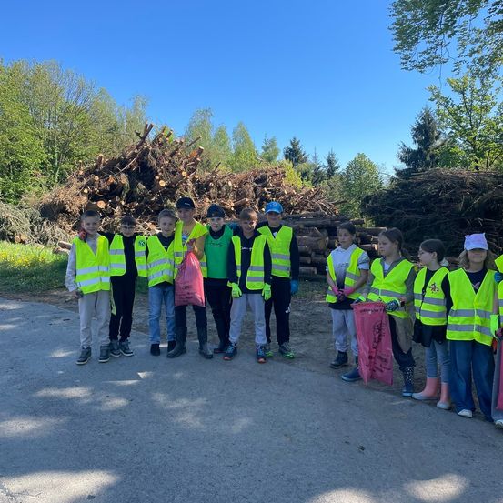 Eine Gruppe junger Menschen in Warnwesten und Handschuhen steht vor einem Holzstapel. Sie haben Plastiktüten und lächeln für ein Foto.