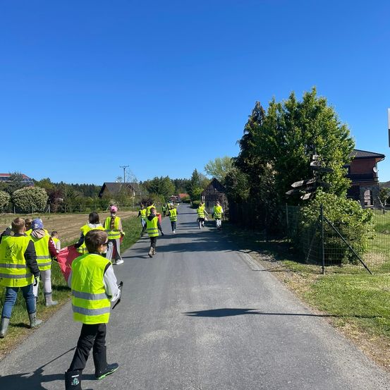 Eine Gruppe von Menschen in neongrünen Westen läuft die Straße entlang. Sie tragen rote Taschen und scheinen auf einer Säuberungsmission zu sein. Bäume und Häuser säumen die Straße.