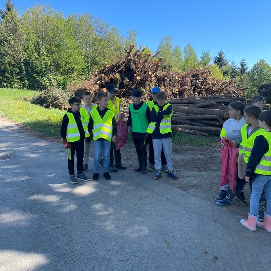 Eine Gruppe von Kindern in Sicherheitswesten und Stiefeln steht auf einem Weg mit einem großen Holzhaufen dahinter.
