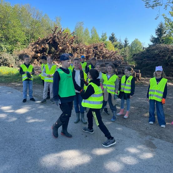 Eine Gruppe von Kindern in Sicherheitswesten steht auf einer Straße, möglicherweise für eine Schulaktivität. Sie stehen in der Nähe eines Holzhaufens.