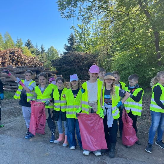 Gruppe junger Menschen in Warnwesten und Handschuhen, die rosa Tüten halten, steht in einem bewaldeten Gebiet.