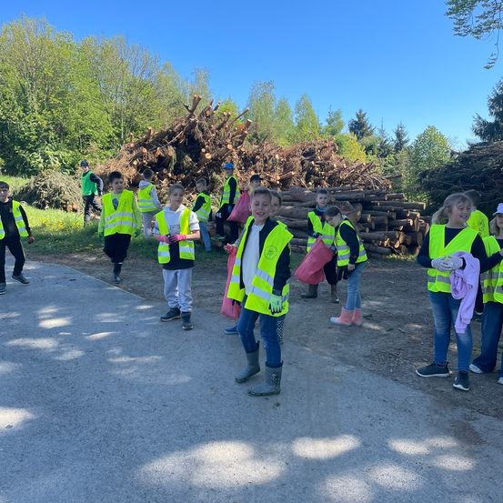 Eine Gruppe von Kindern in Sicherheitswesten und Handschuhen steht auf einer Straße. Dahinter befinden sich Holzhaufen.