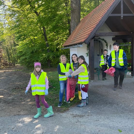 Eine Gruppe junger Freiwilliger in gelben Westen und Stiefeln steht vor einem kleinen Gebäude und hält Plastiktüten in der Hand, bereit für eine Gemeinschaftsreinigung.