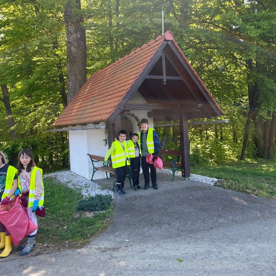 Eine Gruppe von Menschen in gelben Westen und Stiefeln steht vor einer kleinen Kapelle. Bäume umgeben die Kapelle und eine Holzbank steht in der Nähe des Eingangs.