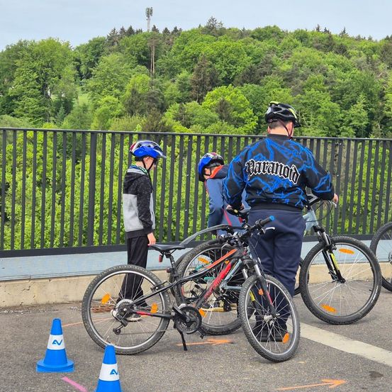Drei Personen bereiten sich auf eine Fahrradtour auf einer asphaltierten Straße mit Verkehrskegel vor. Sie stehen neben ihren Fahrrädern, und einer von ihnen trägt einen blauen Helm.