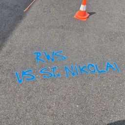 Das Bild zeigt blaue Kreideschrift auf dem Boden, die 'RWS VS ST. NIKOLAI' lautet. Ein orangefarbener Verkehrskegel ist in der Nähe auf der gepflasterten Oberfläche platziert.