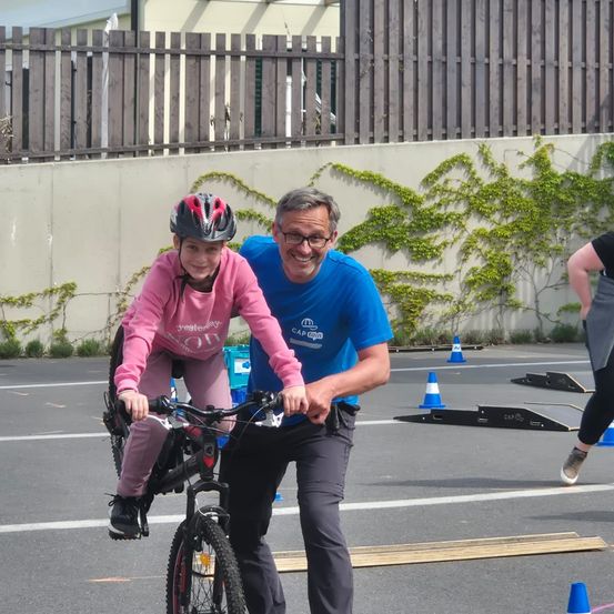 Ein erwachsener Mann führt ein junges Mädchen in einer rosa Jacke auf einem Fahrrad in einem Parkplatz mit einer mit Efeu bedeckten Wand. Eine andere Frau geht im Hintergrund.
