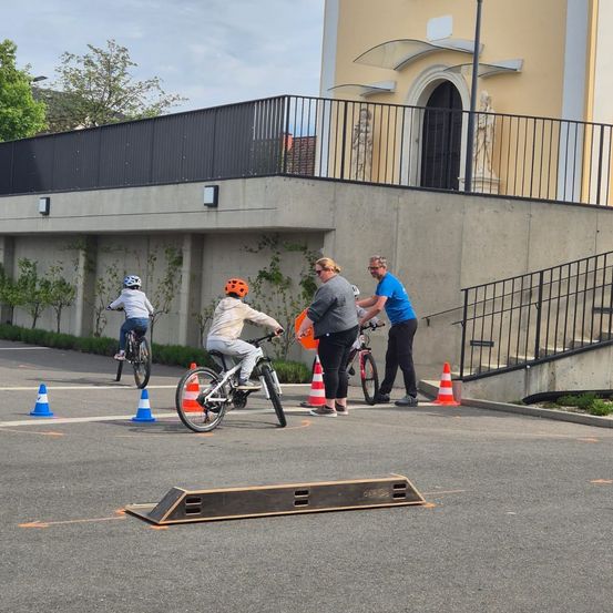 Drei Kinder fahren auf einem Parkplatz mit Fahrrädern, unterstützt von zwei Erwachsenen, umgeben von orangefarbenen Hütchen und einem Gebäude mit Statuen und Treppen.