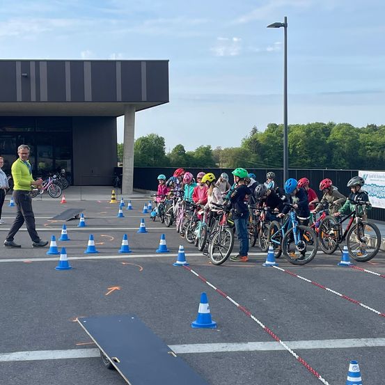 Eine Gruppe von Kindern mit Helmen fährt auf einer von Kegeln umgebenen Strecke mit Fahrrädern, und ein Mann in Gelb steht in der Nähe.