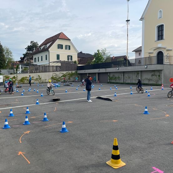 Eine Gruppe von Kindern fährt mit Fahrrädern auf einem Parkplatz, der mit einer durch Kegel markierten Strecke gekennzeichnet ist. Gebäude und Bäume sind im Hintergrund zu sehen.
