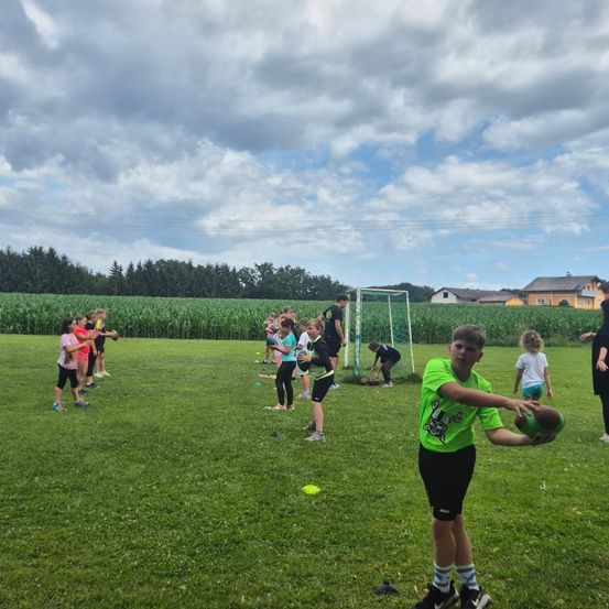 Eine Gruppe von Kindern spielt ein Ballspiel auf einem offenen Feld mit einem Torpfosten im Hintergrund.