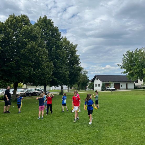 Mehrere Kinder spielen Fangen auf einer Wiese. Ein Mann steht in der Nähe und beobachtet sie. Im Hintergrund befinden sich Bäume und geparkte Autos, ein Haus ist in der Ferne zu sehen.