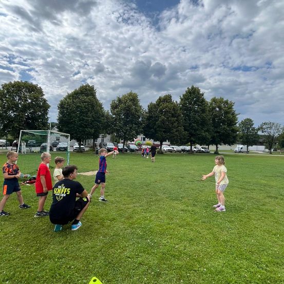 Eine Gruppe von Kindern spielt auf einem Rasenfeld unter einem teilweise bewölkten Himmel. Ein Kind wirft einen Ball, während andere zuschauen. Im Hintergrund steht ein Fußballtor und mehrere Autos sind geparkt.