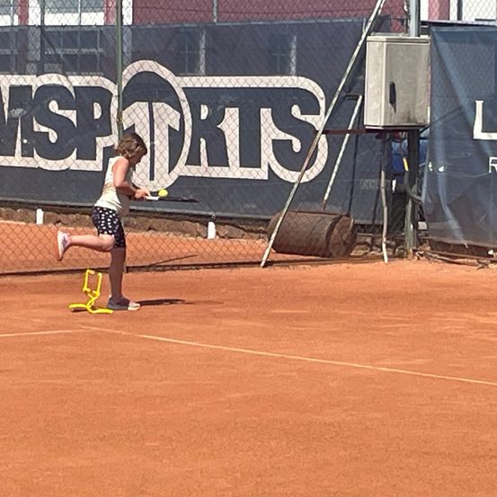 Ein junges Mädchen übt Tennis auf einem Sandplatz. Sie schlägt einen gelben Tennisball. Hinter ihr befindet sich ein Zaun mit einem großen Banner, das MSP Sports bewirbt.