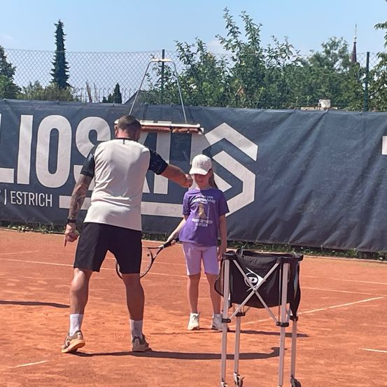 Ein Tennistrainer unterrichtet ein junges Mädchen auf einem Tennisplatz mit einem Banner im Hintergrund.