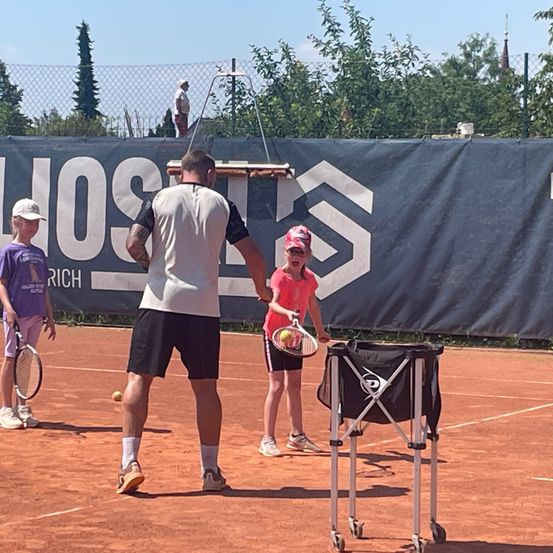 Ein Tennistrainer unterrichtet zwei Kinder auf einem Sandplatz, mit einem Tennisballhalter in der Nähe und einem Banner im Hintergrund.