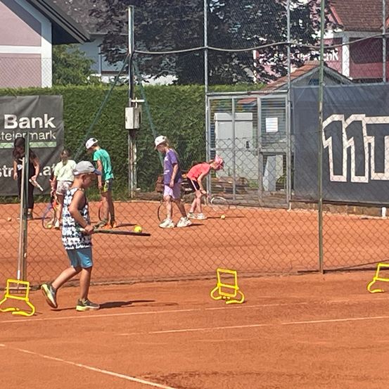 Eine Gruppe von Kindern spielt Tennis auf einem Sandplatz. Einige beugen sich vor, während andere Tennisschläger halten. Hinter ihnen steht ein Zaun mit einem Banner.