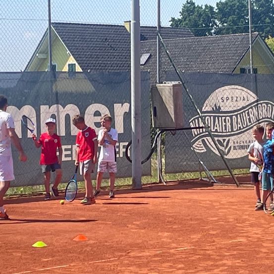 Ein Tennislehrer unterrichtet Kinder auf einem Sandplatz, drei Jungen halten Schläger, ein weiterer Junge geht, und ein Erwachsener geht im Vordergrund.